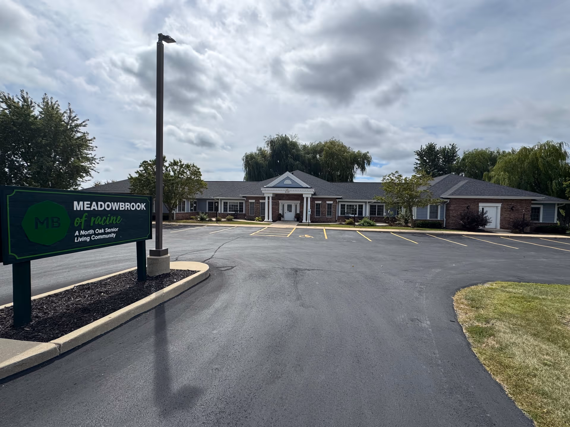 Front exterior of Meadowbrook of Racine senior living building with entrance, parking lot, and property sign in the foreground.