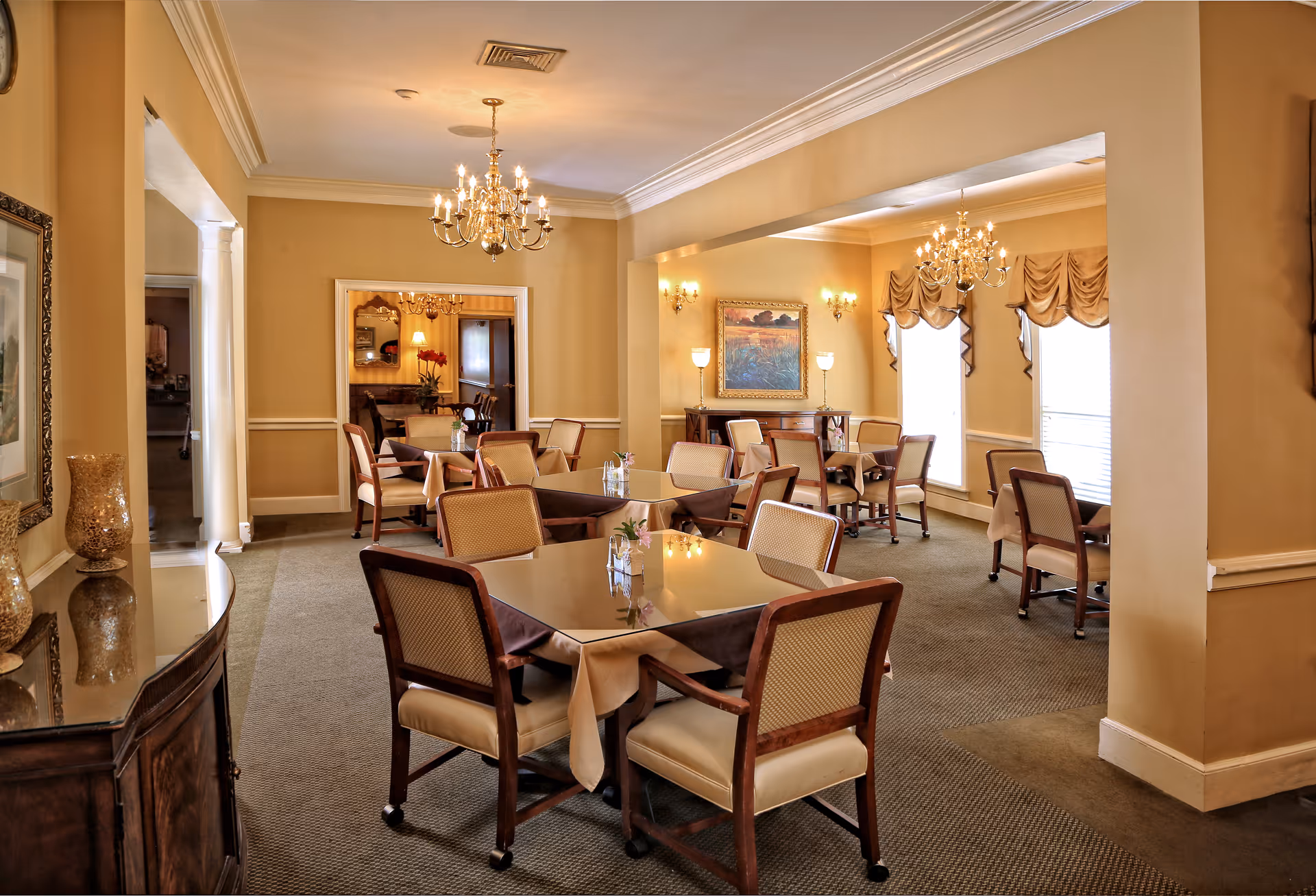 Elegant senior living dining room with multiple set tables, upholstered chairs, chandeliers, and warm beige decor.