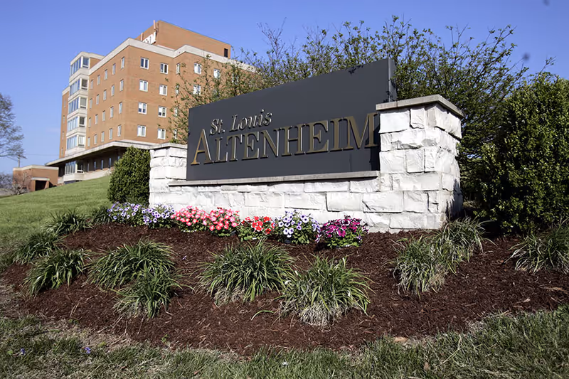 Stone entrance sign reading 'St. Louis Altenheim' with a flowerbed in front and the facility building visible behind it.