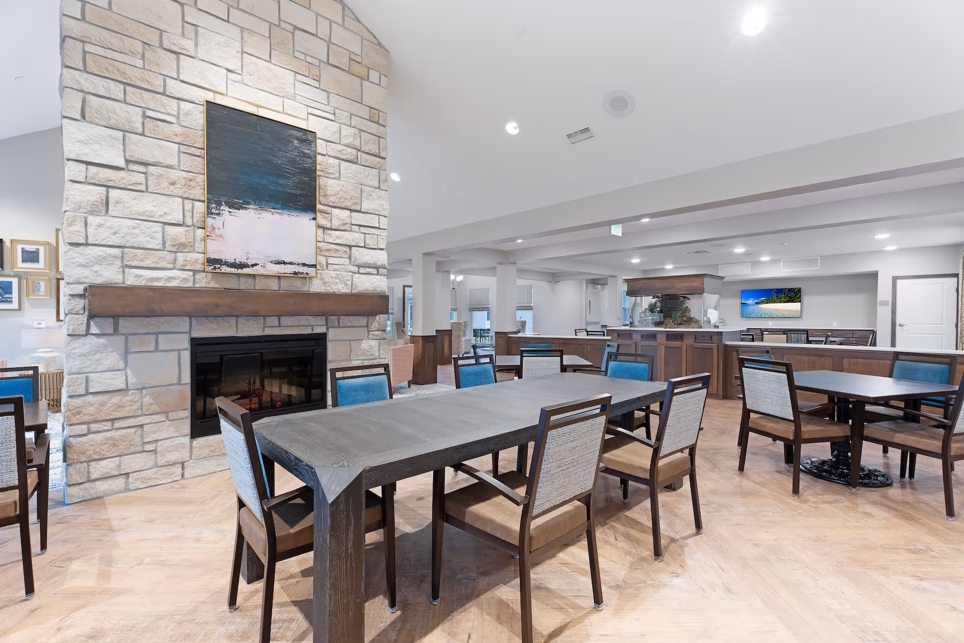 A spacious dining area in a senior living facility featuring multiple tables and chairs arranged neatly. The room has a large stone fireplace with a modern abstract painting above it. The flooring is light wood, and the walls are painted in neutral tones. In the background, there is a counter area with a mounted flat-screen TV displaying a beach scene.