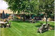 Outdoor garden area at Hickory House Nursing Home with green grass, several wooden benches and picnic tables, large trees providing shade, and umbrellas over some tables. The building is visible in the background.