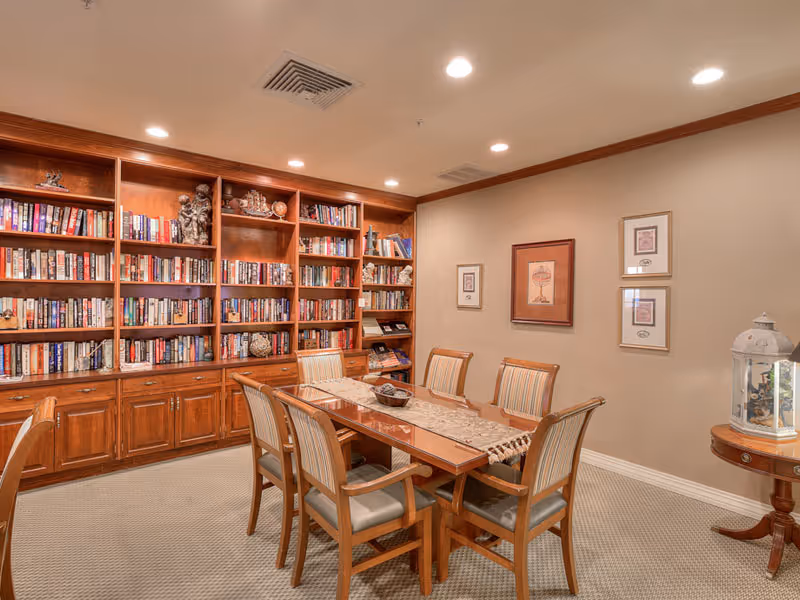 A cozy room with a wooden table surrounded by six cushioned chairs. Behind the table is a large wooden bookshelf filled with books and decorative items. The walls are beige with three framed pictures hanging, and a small round table with a decorative lantern is placed in the corner. The room is well-lit with recessed ceiling lights.