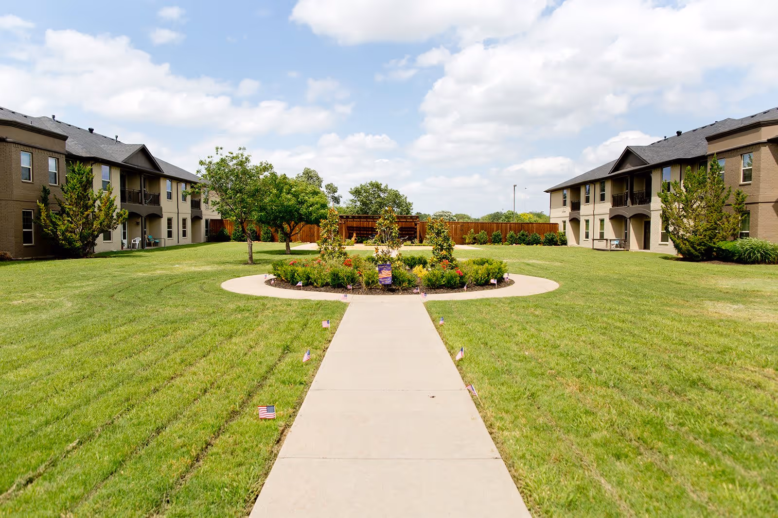A wide concrete pathway leads through a well-maintained grassy courtyard with a circular flower bed in the center. The courtyard is surrounded by two-story beige residential buildings with balconies and windows. Small American flags line the pathway, and there are trees and shrubs around the area under a partly cloudy sky.