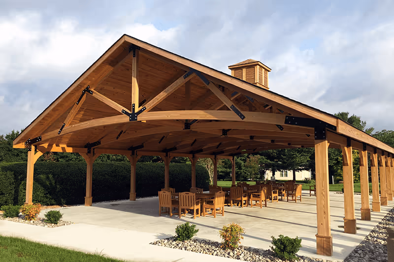 A large wooden outdoor pavilion with exposed beams and several tables and chairs arranged on a concrete patio.