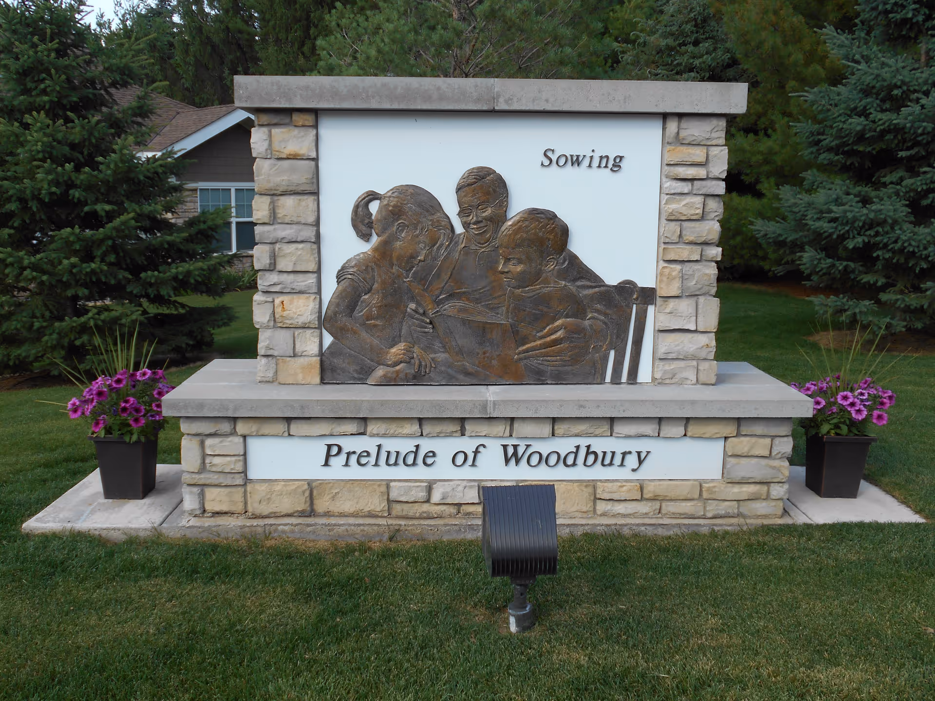 Stone monument sign with a bronze relief sculpture of three people reading a book together, labeled 'Sowing'. The base of the monument reads 'Prelude of Woodbury'. The sign is flanked by two planters with purple flowers and surrounded by green grass and trees.