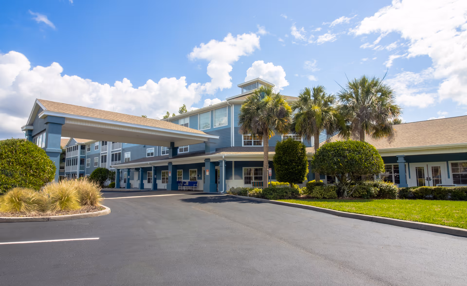 Exterior view of Addington Place of East Lake, a multi-story senior living facility with blue siding and beige roof. The entrance features a covered drop-off area with columns. There are palm trees, trimmed bushes, and landscaped grass around the building under a partly cloudy blue sky.