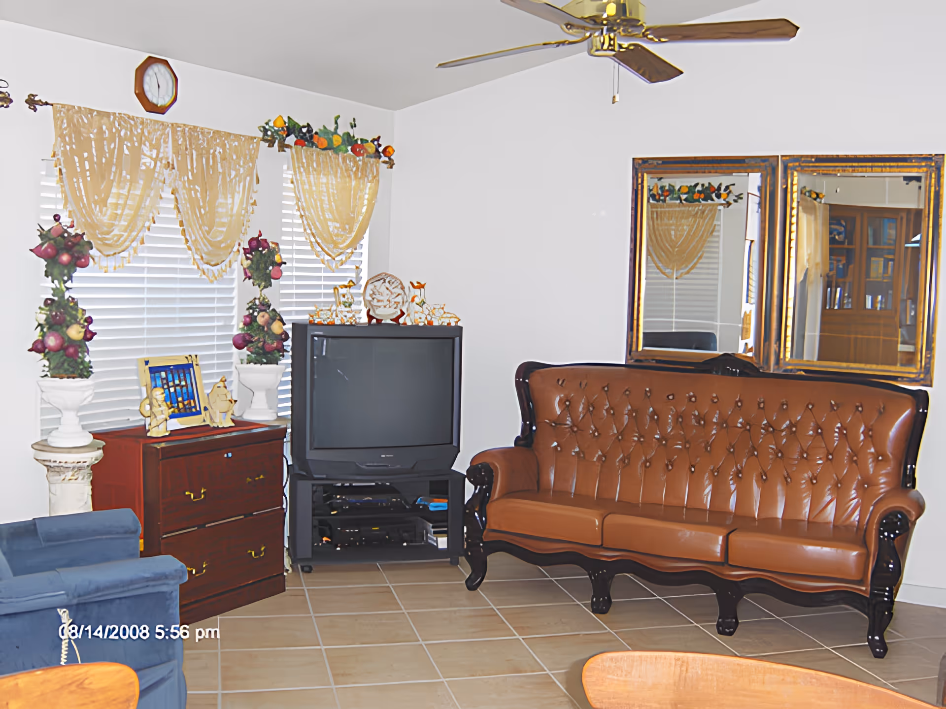 A living room with a brown leather tufted sofa, a blue armchair, a wooden cabinet with decorative items, a TV on a black stand, two large mirrors on the wall, and windows with yellow curtains and floral decorations. The floor is tiled, and there is a ceiling fan above.