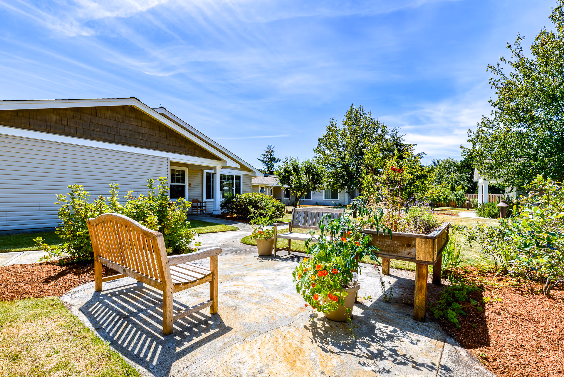 Sunny courtyard with wooden benches, planter boxes, and landscaping in front of a single-story memory care building.