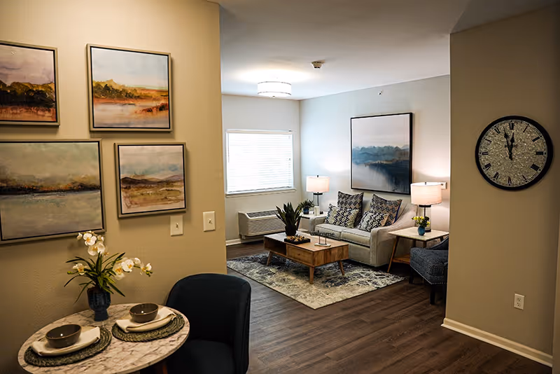A cozy senior living room with a small round dining table set for two with bowls and plates, a vase with white flowers, and a dark blue chair. The living area features a light gray sofa with patterned cushions, a wooden coffee table with a plant, two side tables with lamps, a large wall clock, and framed landscape paintings on the walls. The room has wood flooring and a window with blinds.
