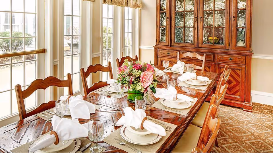 A well-lit dining room with a wooden dining table set for six people. The table has white plates, folded white napkins in wooden rings, clear drinking glasses, and silverware. A floral centerpiece with pink and red flowers is placed in the middle of the table. Behind the table is a wooden china cabinet with glass doors displaying dishes. Large windows with white frames and beige curtains allow natural light to fill the room.