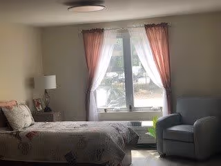 A bright bedroom with a single bed, nightstand and lamp, an armchair, and a window with pink curtains.