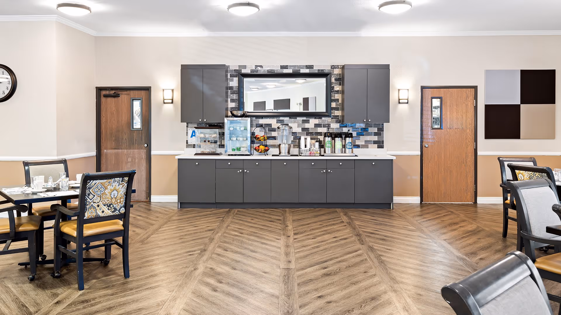 A clean and well-lit dining area with wooden flooring and beige walls. There are several tables with chairs arranged around them, some set with plates and cups. Against the far wall is a counter with dark gray cabinets, a large mirror, a water dispenser, coffee machines, and a fruit basket. Two wooden doors flank the counter, and a clock is visible on the left wall.