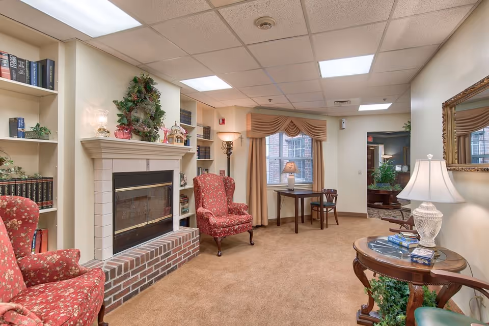 Cozy assisted living lounge with a fireplace, upholstered armchairs, bookshelves, and table lamps.