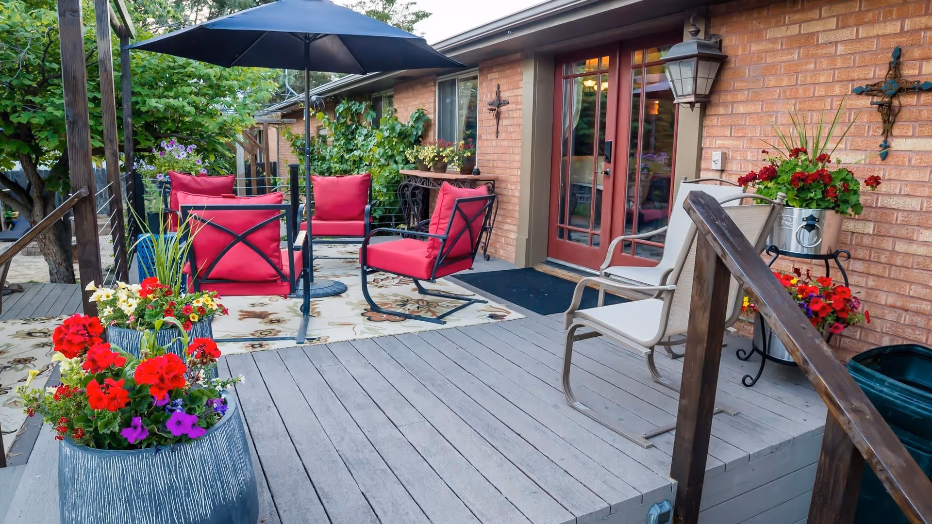 Outdoor patio area with red cushioned chairs, a large umbrella, potted flowers, and a brick wall with red-framed glass doors leading inside. The patio has wooden flooring and decorative plants around.