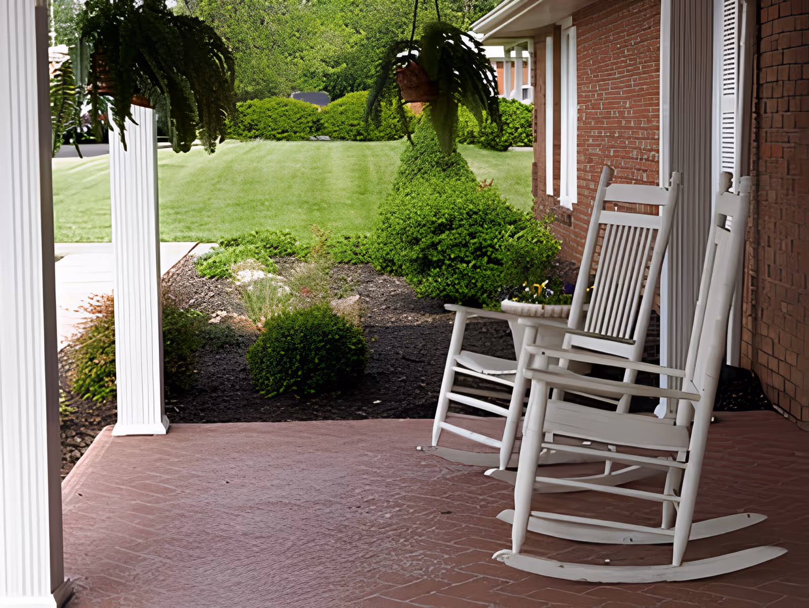 Covered front porch with white rocking chairs, hanging ferns, and a landscaped lawn beyond.