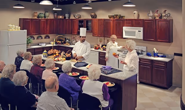 A chef and two staff demonstrate cooking in a community kitchen to a seated group of senior residents.