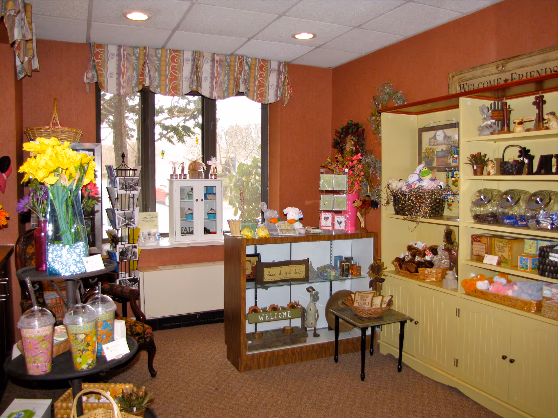 Interior view of a small gift shop area with shelves and display cases filled with various decorative items, stuffed animals, and small gift products. There is a window with patterned valance curtains, a small table with baskets, and a vase with yellow flowers on a stand.