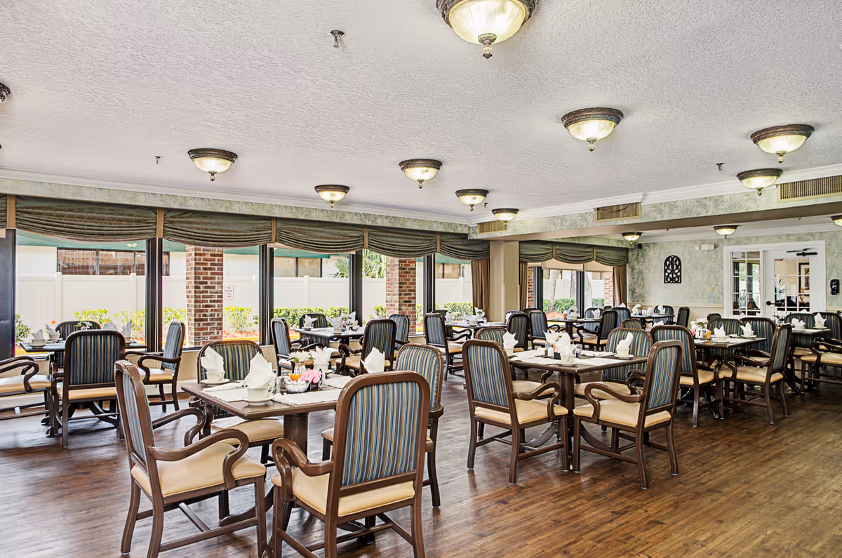 Spacious dining room with multiple set tables and upholstered chairs, hardwood floors, ceiling lights, and large windows along one wall.
