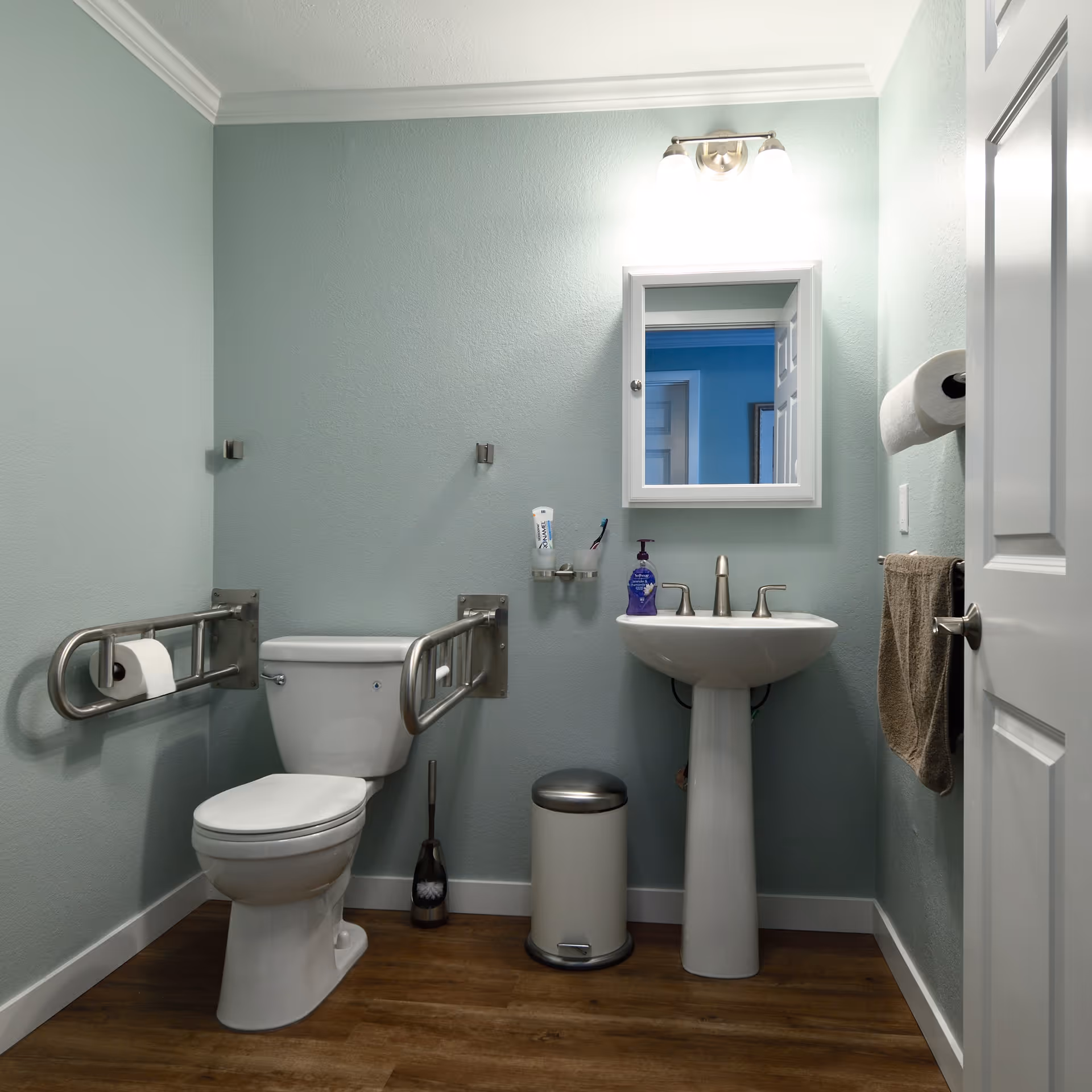 A clean and accessible bathroom with light blue walls, a white toilet equipped with metal grab bars on both sides, a white pedestal sink with a mirror above it, a wall-mounted light fixture, a towel hanging on the wall, a trash can, and a toilet brush on the wooden floor.