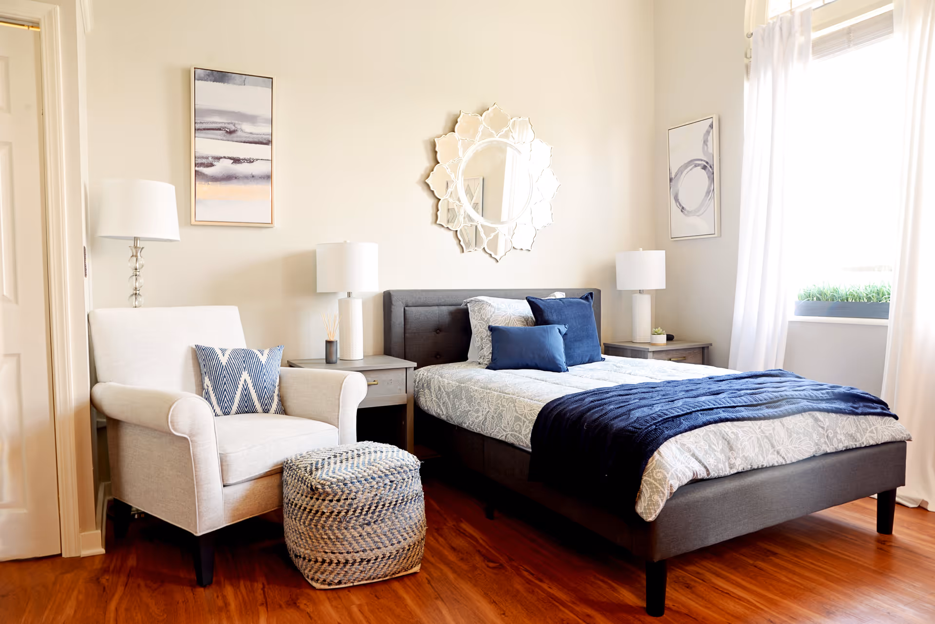 A bright and cozy bedroom with a gray upholstered bed featuring white and blue bedding, two matching nightstands with lamps, a white armchair with a blue patterned pillow, a woven ottoman, a decorative mirror above the bed, and two abstract paintings on the walls. Sunlight streams in through a window with white curtains.