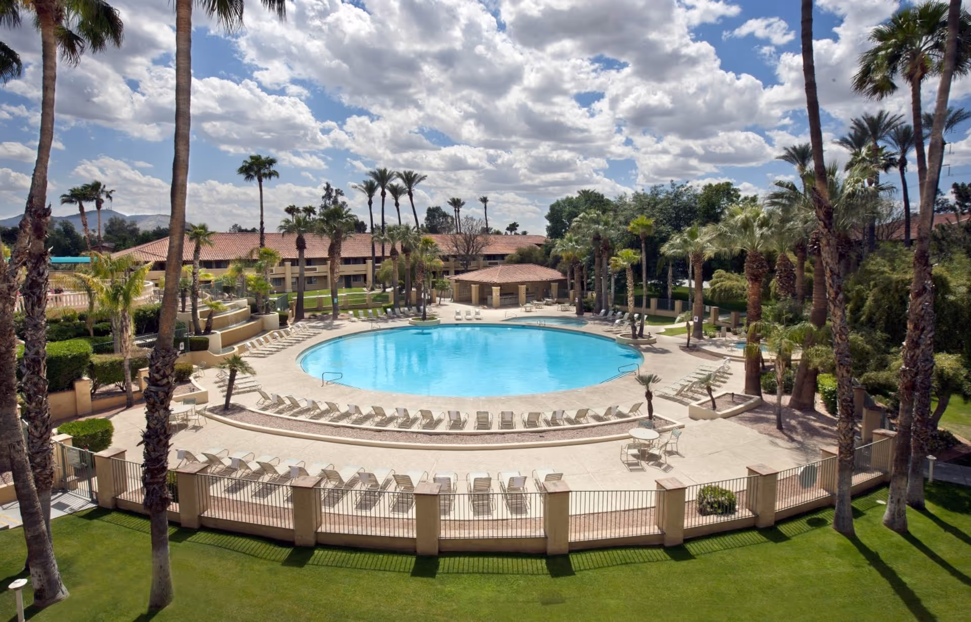 Large circular outdoor swimming pool surrounded by lounge chairs and palm trees on the grounds of a senior living facility.
