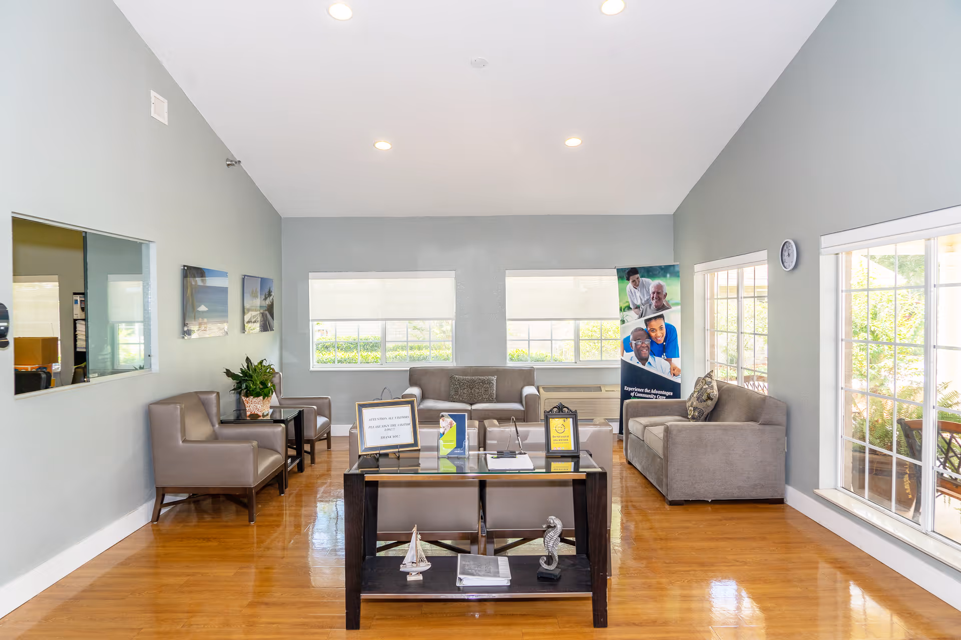 A bright and clean living room area in a nursing home with gray walls and wooden flooring. The room features two gray sofas, two gray armchairs, a glass coffee table with decorative items, and large windows letting in natural light. There are framed pictures on the wall and a standing banner with images of smiling elderly people and caregivers.