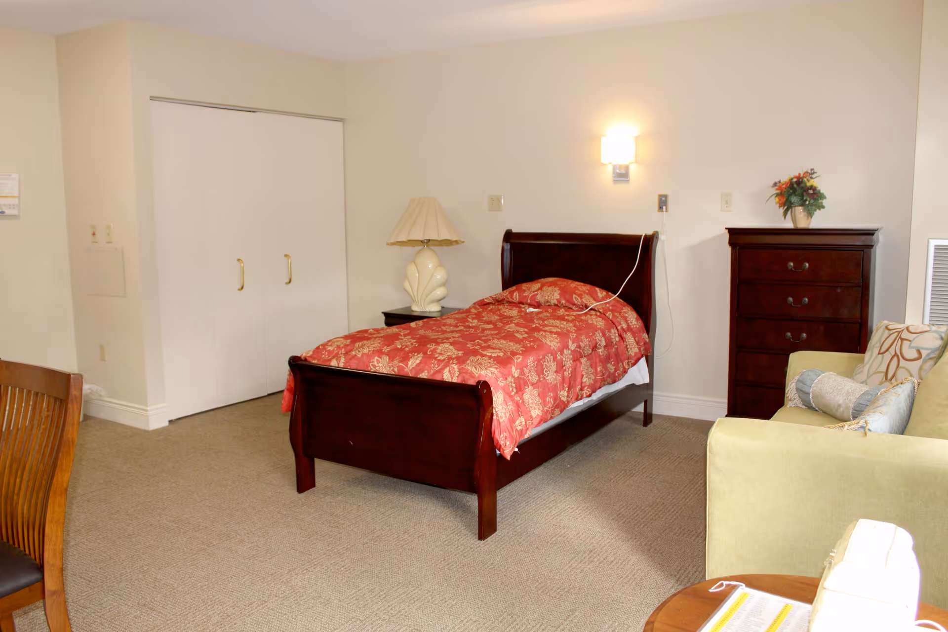 A furnished assisted-living bedroom with a single wooden bed with a red floral bedspread, nightstand and dresser, and a small sofa.