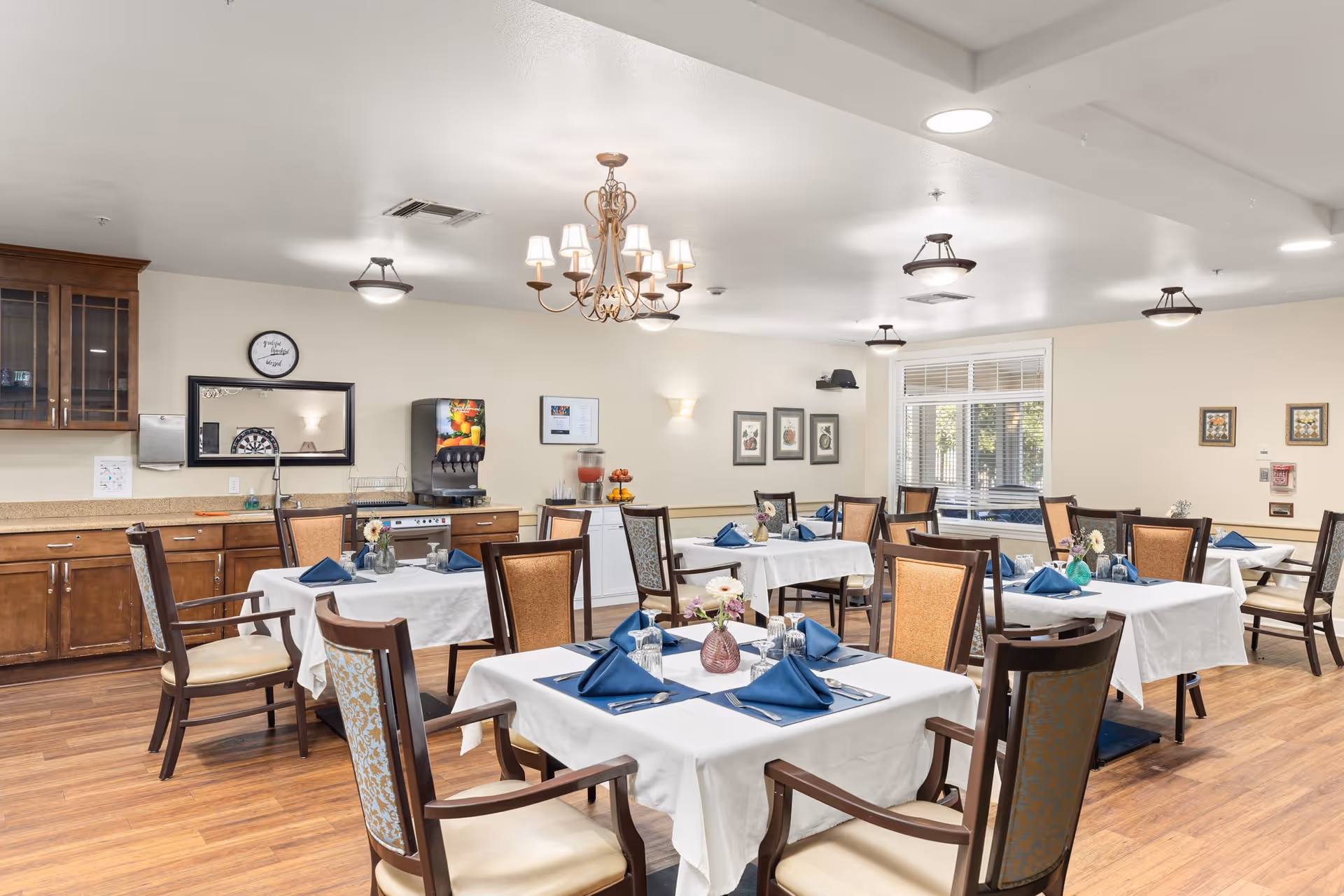 A bright dining room with several tables covered in white tablecloths, each set with blue napkins, silverware, and small flower vases. The room features wooden chairs with cushioned seats and backs, wood flooring, and a beverage dispenser against the far wall. There are framed pictures on the walls and a window letting in natural light.