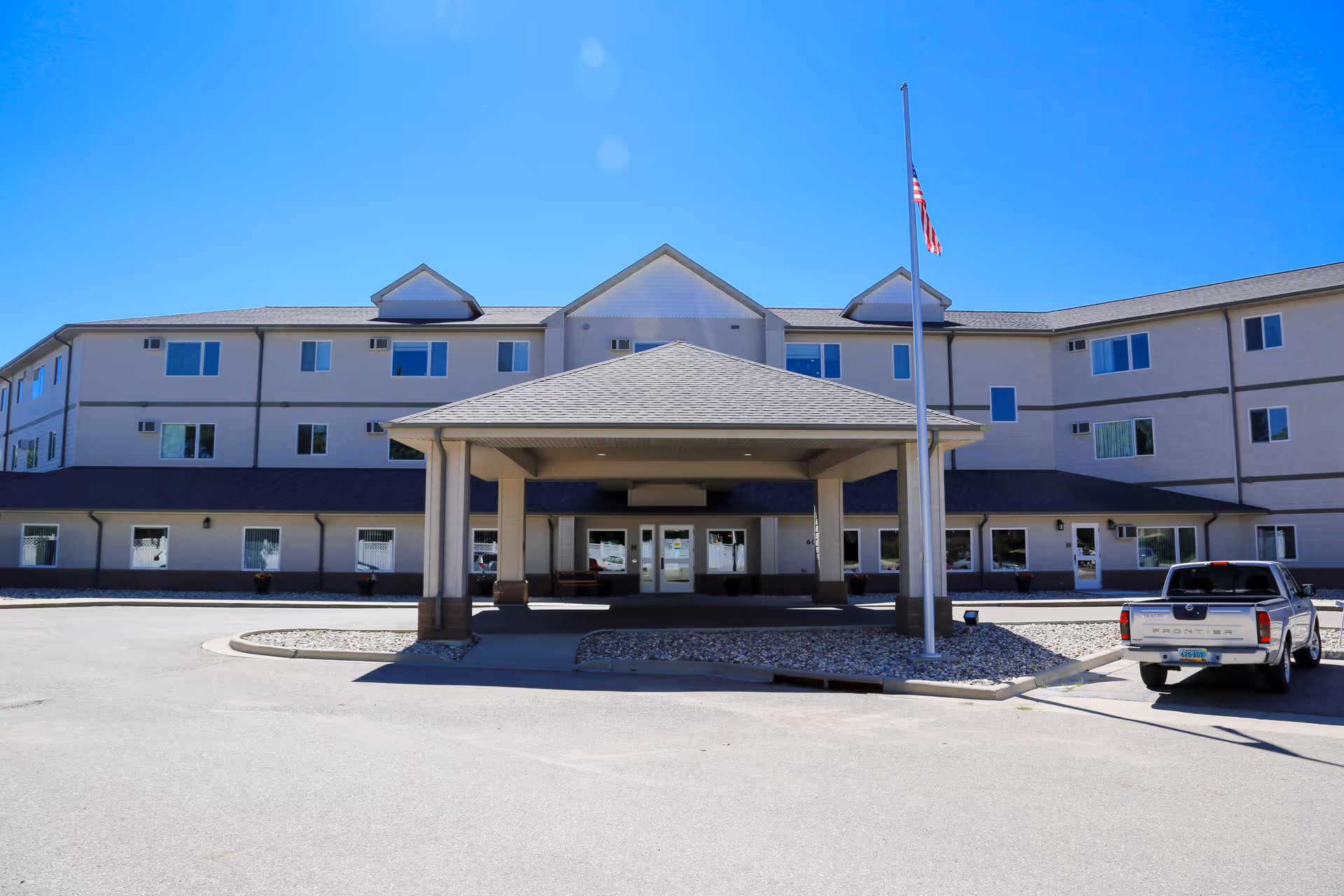 Exterior front view of a three-story senior living facility building with a covered entrance, an American flag on a flagpole, and a parked pickup truck in the driveway under a clear blue sky.