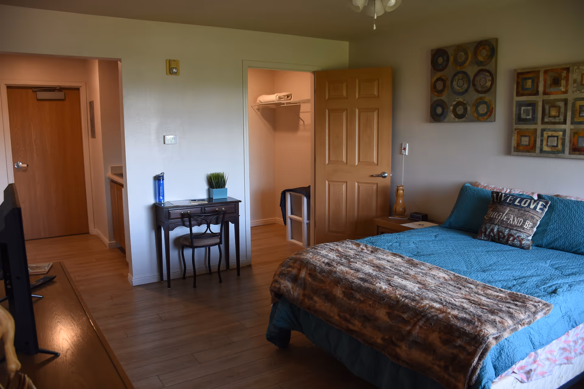 A cozy bedroom in an assisted living community featuring a bed with blue bedding and a brown throw blanket, a decorative pillow with text, two pieces of wall art above the bed, a wooden nightstand with a lamp, a small desk with a chair and a plant, an open door revealing a closet with towels, and a hallway leading to another door.