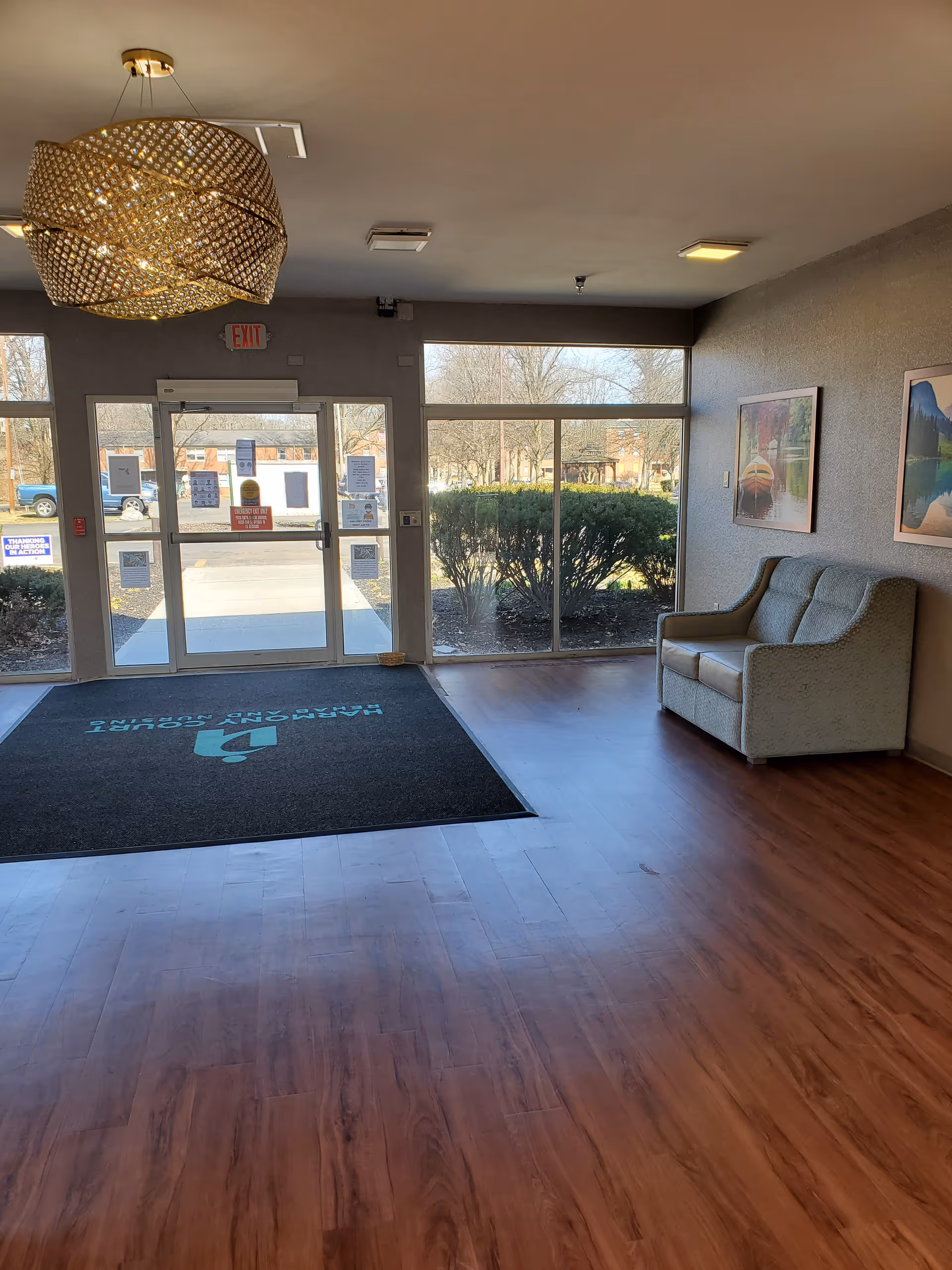 Interior view of a facility entrance area with wooden flooring, a large decorative ceiling light fixture, a glass door with exit sign above it, large windows showing bushes and trees outside, a black floor mat with facility name, and a two-seater couch against the right wall with framed pictures above it.