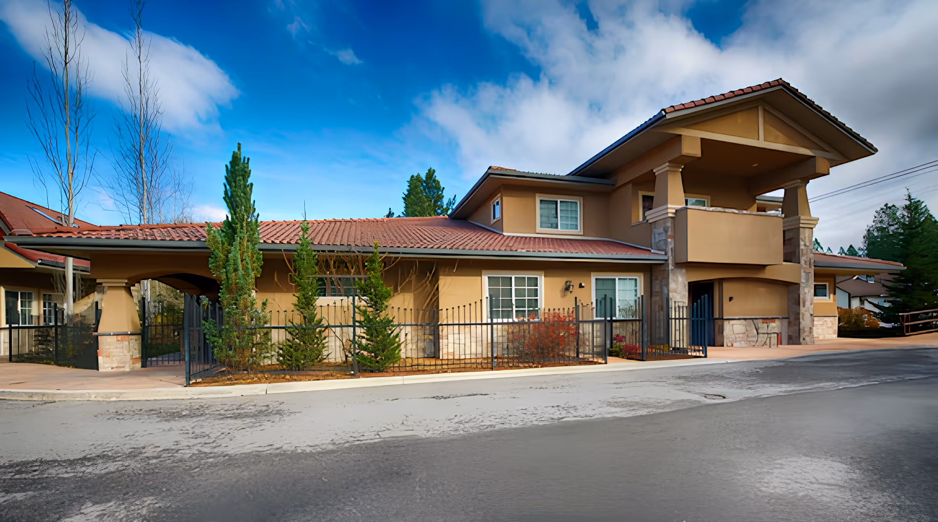 Exterior view of a senior living facility building with beige walls, red tiled roof, and a gated entrance. The building has a covered balcony and is surrounded by small trees and shrubs under a partly cloudy blue sky.