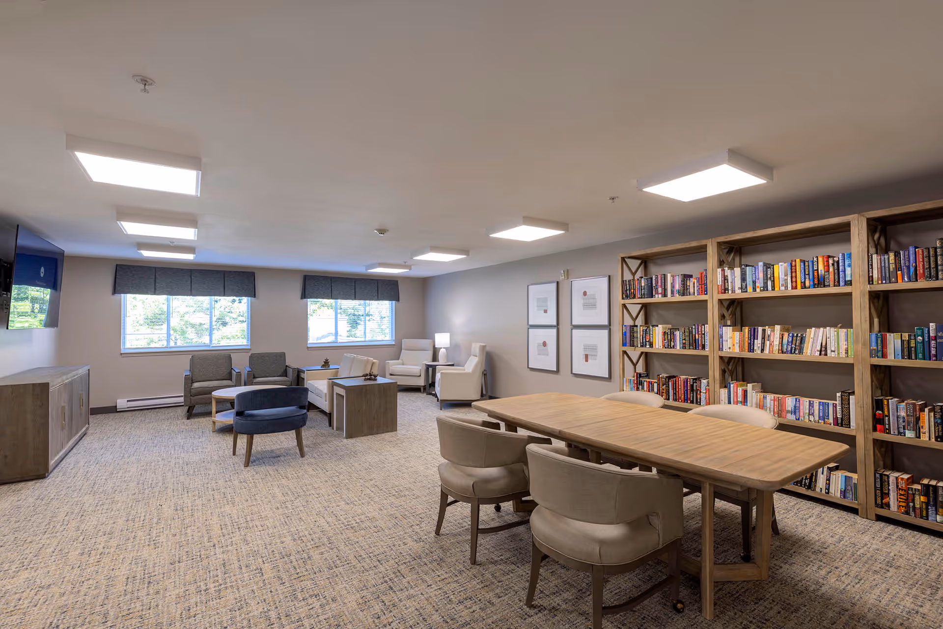 A spacious, well-lit common room with a large wooden table surrounded by six upholstered chairs in the foreground. Behind the table is a large bookshelf filled with books. In the background, there are several armchairs and a sofa arranged around a coffee table near two windows with gray valances. A flat-screen TV is mounted on the wall to the left, above a wooden cabinet. The room has a neutral color palette with beige walls and carpeted flooring.
