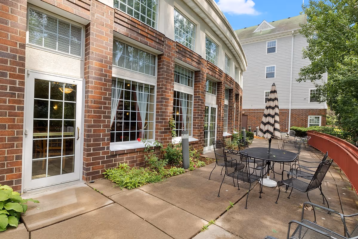 Outdoor patio area at Eagle Crest Assisted Living featuring a brick building with large windows and glass doors. The patio has metal tables and chairs with a closed striped umbrella, surrounded by greenery and a red railing.