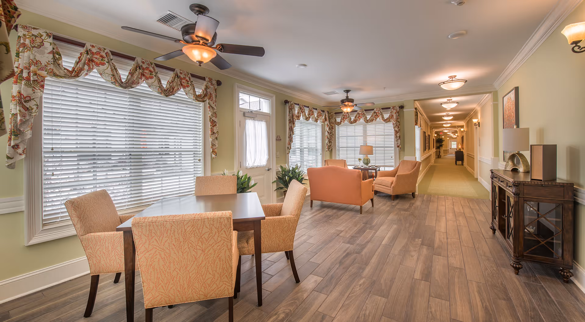 A bright and spacious interior common area with large windows covered by floral valances and white blinds. The room features a wooden table with four upholstered chairs on the left, and a seating area with two armchairs and a small table with a lamp near the windows. The floor is covered with wood-like tiles, and a long hallway extends into the background. The walls are painted light green with white trim, and ceiling fans with lights are mounted on the ceiling.