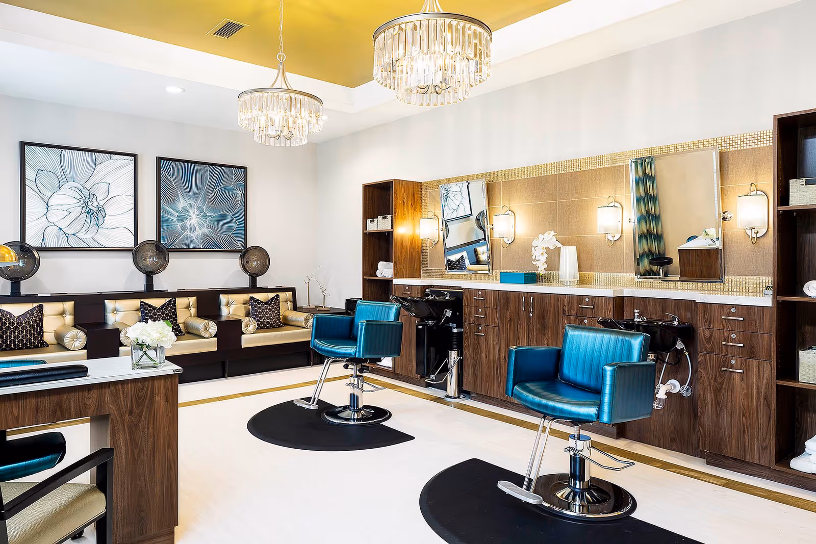A modern hair salon area with two teal salon chairs in front of a long counter with sinks and mirrors. The room features wooden cabinetry, two chandeliers, and a seating area with beige cushioned benches and decorative pillows. Two large floral artworks hang on the wall above the seating area.