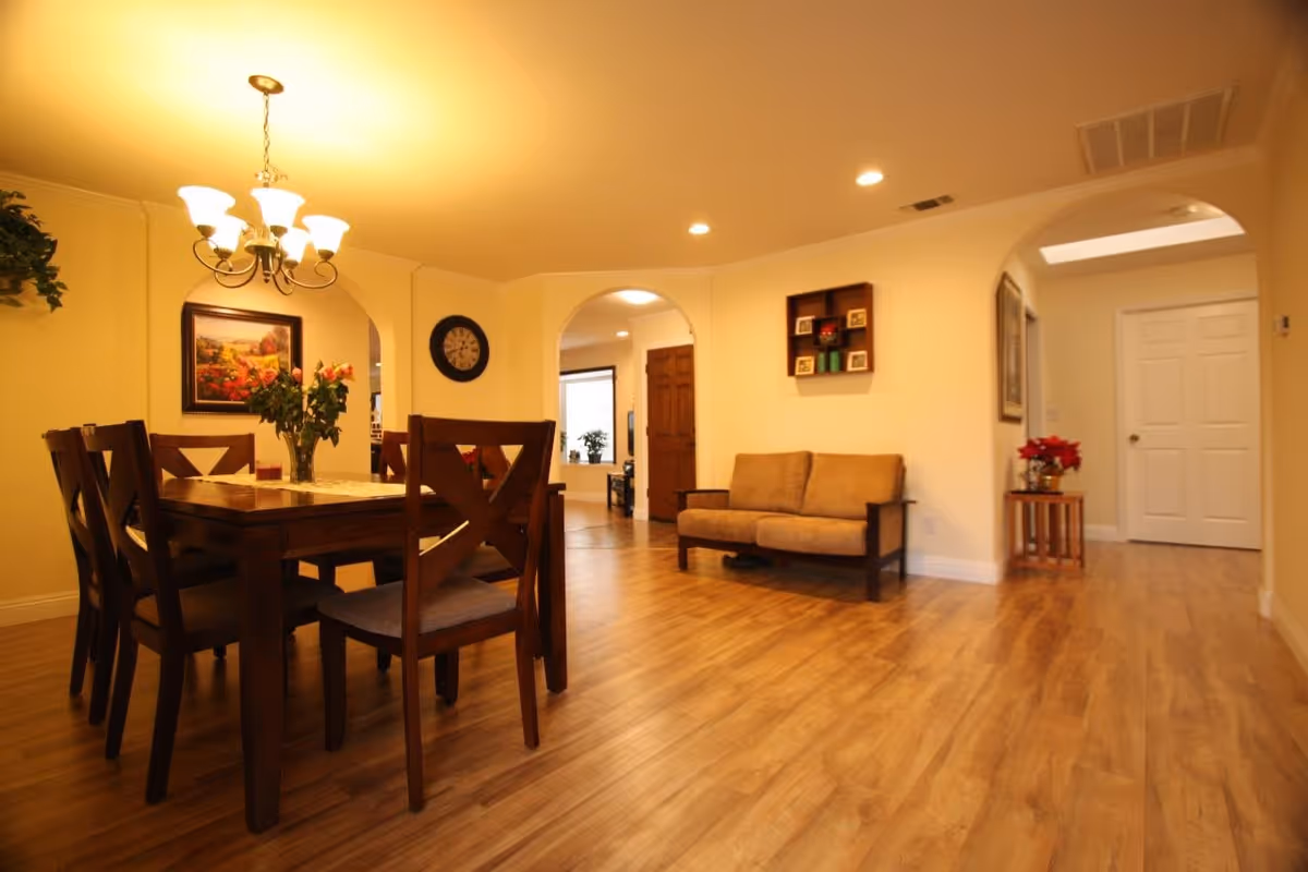 A warmly lit interior space featuring a wooden dining table with six chairs and a vase of flowers on top. To the right, there is a small beige couch against the wall with a decorative shelf above it. The room has wooden flooring, cream-colored walls, and arched doorways leading to other parts of the facility. A chandelier hangs above the dining table, and a clock and framed artwork are visible on the walls.