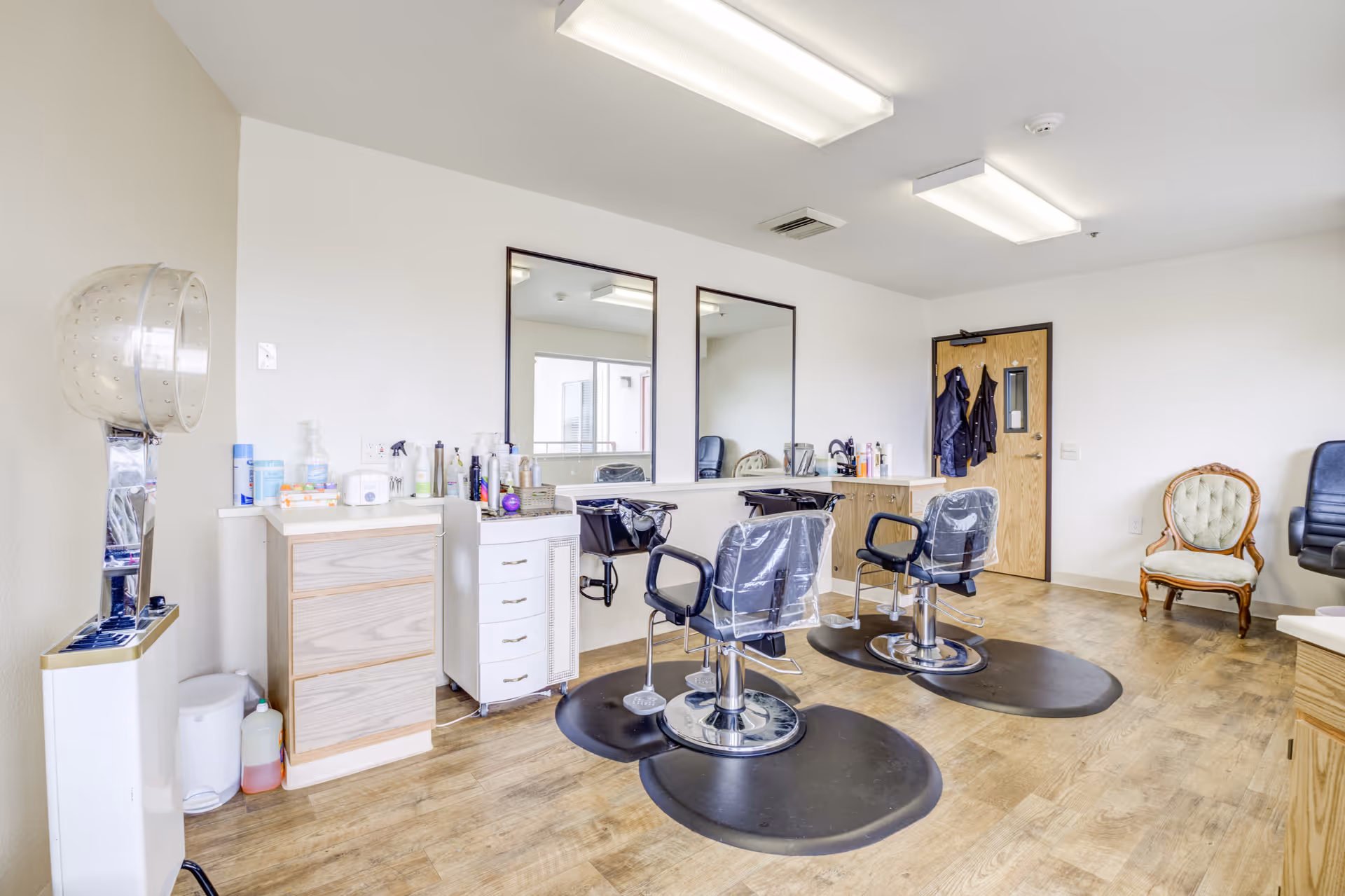 A bright and clean salon area with two black salon chairs covered in protective plastic, positioned in front of a long counter with two large mirrors. The room has wooden flooring, a wooden door with coats hanging on hooks, a vintage upholstered chair, and a hair dryer station on the left side.