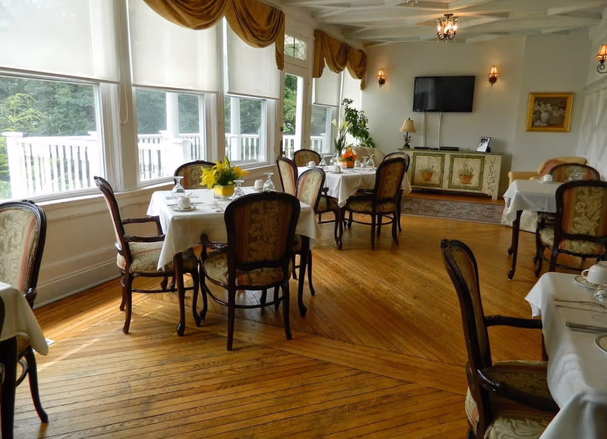 Bright dining room with round tables set with white tablecloths and floral centerpieces, upholstered chairs, large windows and hardwood floors.