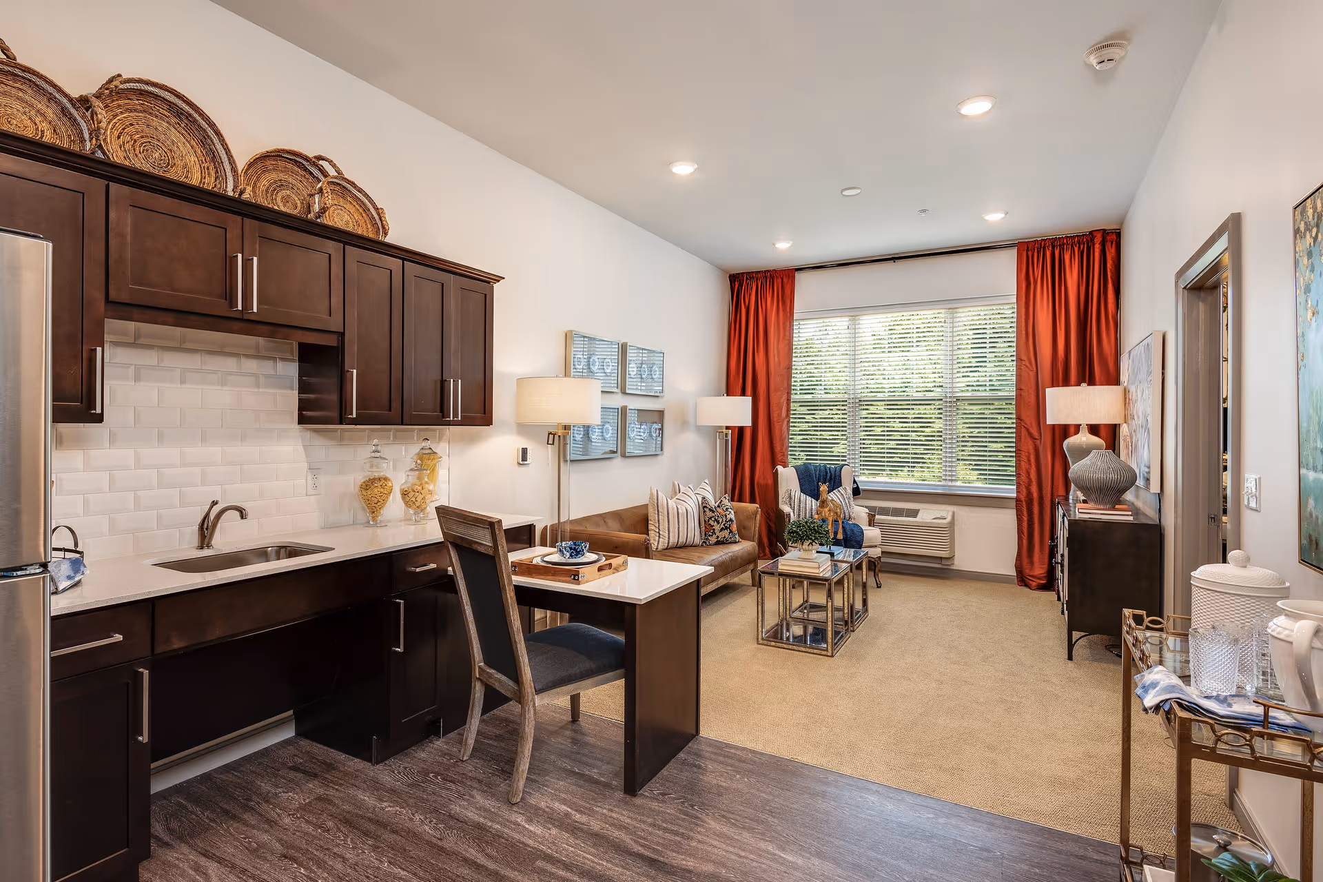 A well-lit interior living space featuring a small kitchen area with dark wood cabinets, a sink, and a refrigerator on the left. Adjacent to the kitchen is a dining table with a chair. The living room area has a brown sofa with decorative pillows, a glass coffee table, two armchairs, and floor lamps. Large windows with red curtains allow natural light to fill the room. The floor transitions from wood in the kitchen area to carpet in the living area.