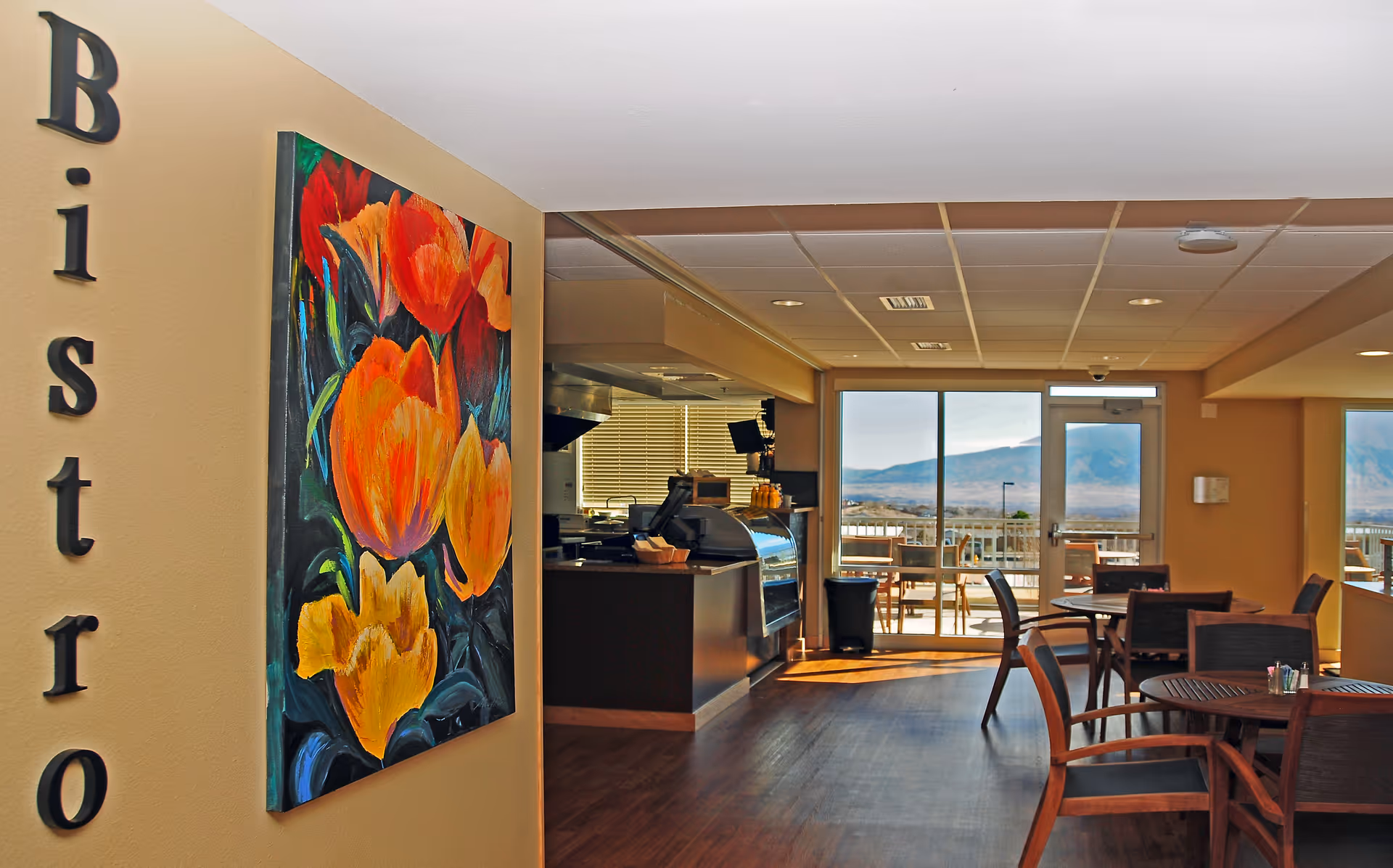 Interior view of a bistro area with wooden floors, several round tables with chairs, a counter with a display case, and large windows showing an outdoor patio and distant mountains. A colorful painting of orange and red flowers is mounted on a wall with the word 'Bistro' displayed vertically.