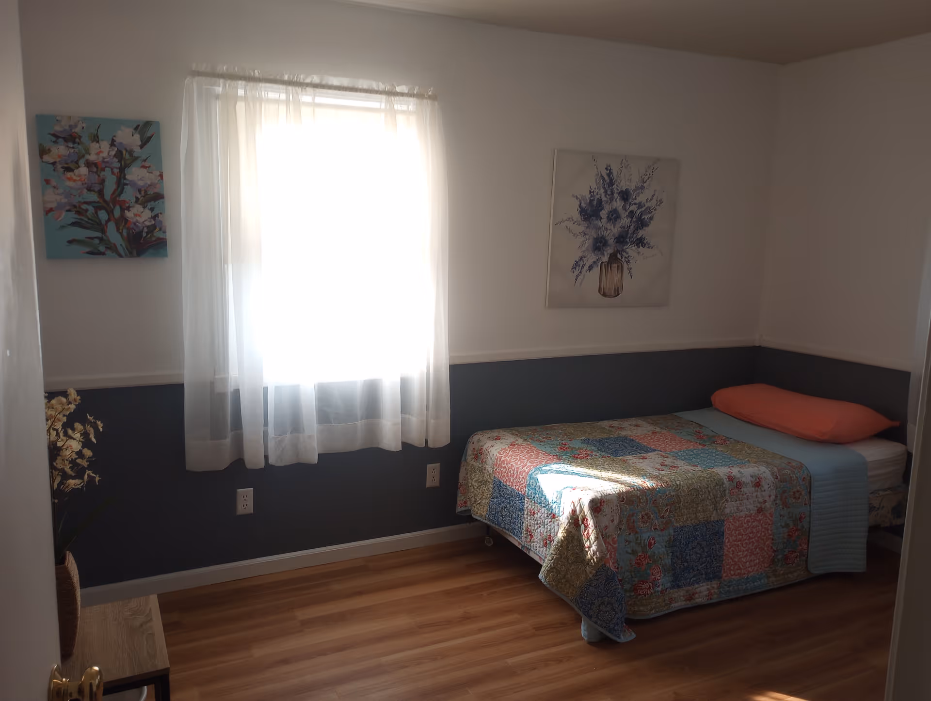 Small bedroom with a single bed covered by a patchwork quilt, a window with sheer curtains, and floral wall art.
