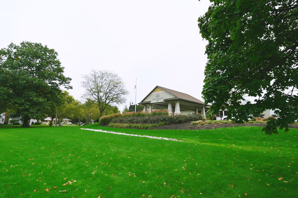A lush green lawn with scattered fallen leaves and a small white rock pathway leading to a covered pavilion structure labeled 'Vista Springs'. The area is surrounded by large trees with green foliage under an overcast sky.