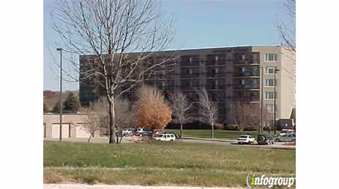 Exterior view of a multi-story senior living facility building with several balconies, a parking lot with cars, leafless trees, and a grassy area in the foreground under a clear sky.