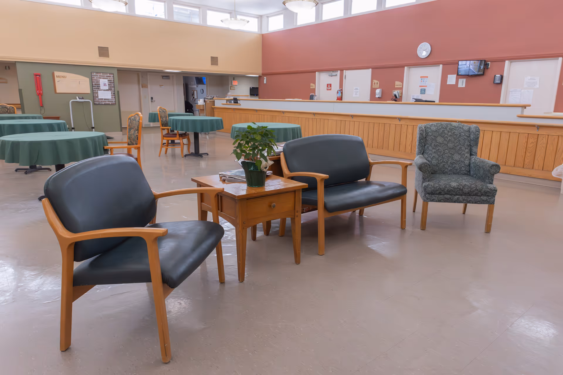 A spacious common area in a senior living facility with several green-covered round tables and wooden chairs in the background. In the foreground, there are two black cushioned wooden armchairs, a matching loveseat, and a patterned armchair arranged around a small wooden table with a potted plant on it. The walls are painted in warm tones with a clock and a TV mounted on the far wall.