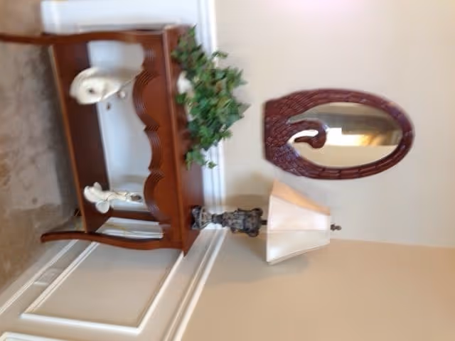 A wooden console table against a beige wall with decorative items including a white ceramic swan, a green leafy plant, and a small statue. Above the table is an oval mirror with a dark wooden frame. A table lamp with a beige shade is placed on the right side of the table.