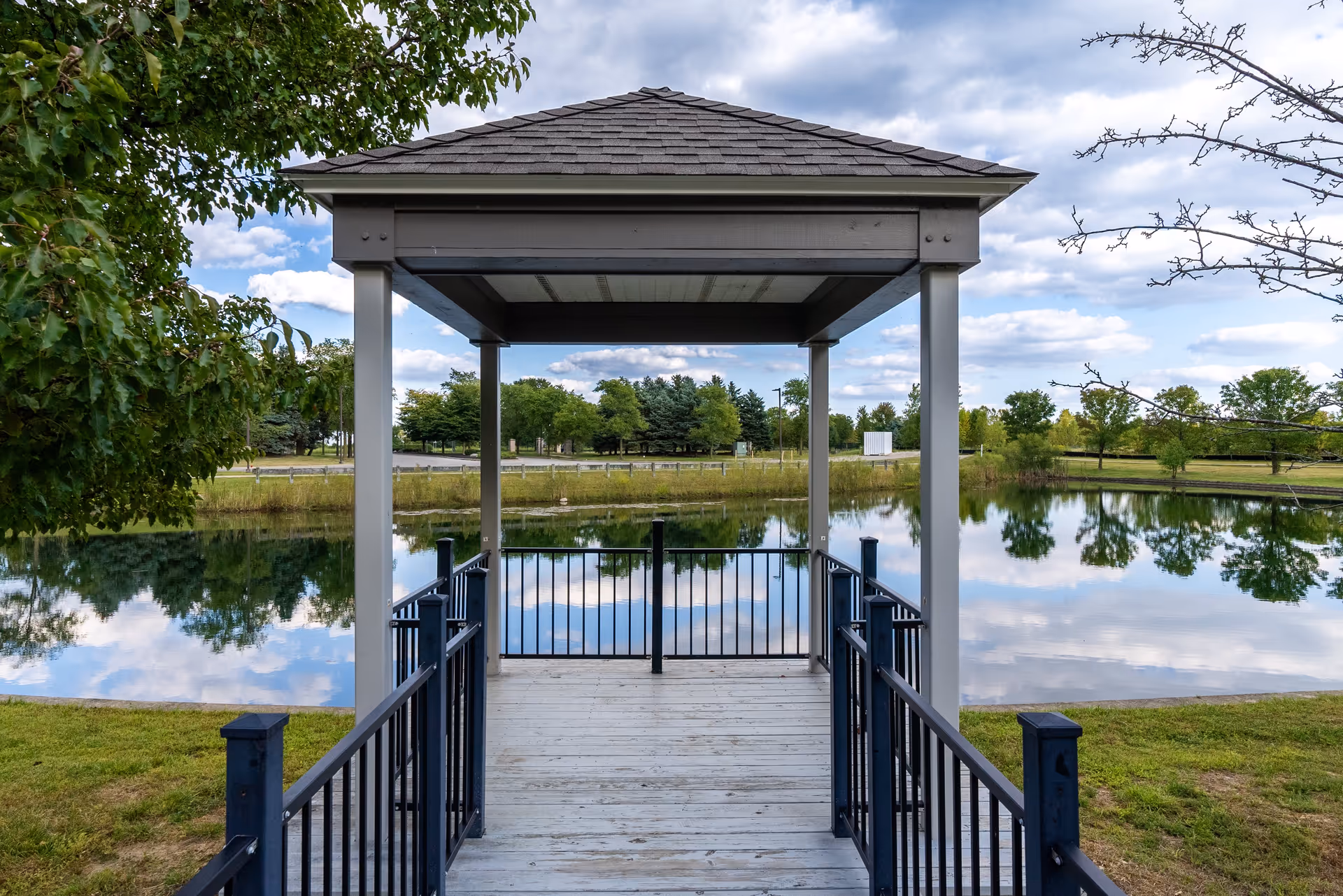 A wooden gazebo with a shingled roof and black metal railings overlooking a calm pond surrounded by green grass and trees under a partly cloudy sky.