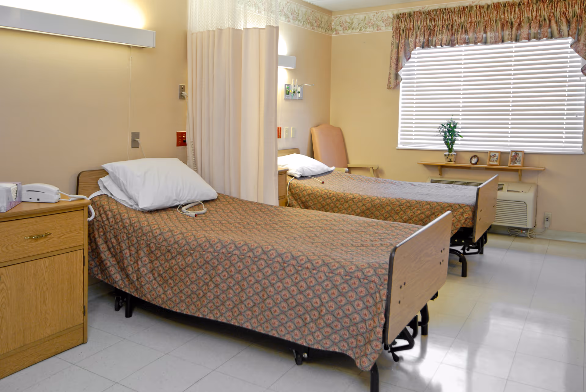Two hospital-style beds with patterned bedspreads in a shared senior living room with a window, nightstand, and privacy curtain.