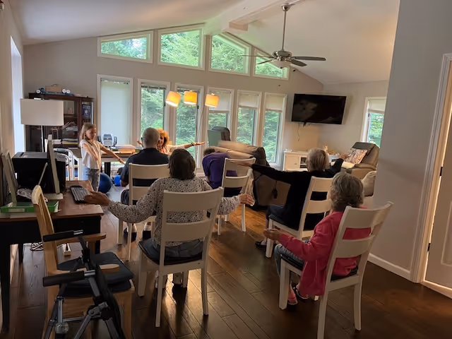 A group of elderly people seated on chairs in a living room, participating in a seated exercise session led by a young girl standing in front of them. The room has large windows with natural light, a ceiling fan, a wall-mounted TV, and comfortable furniture including sofas and a bookshelf.