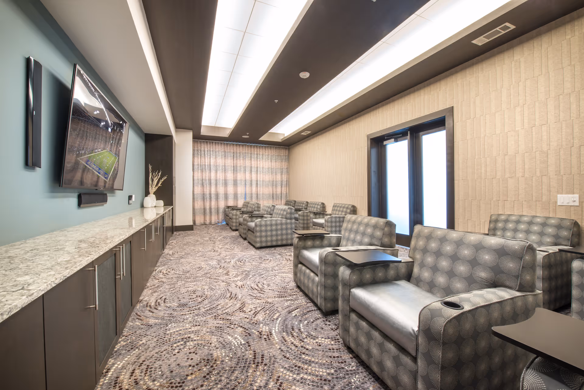 A modern lounge or media room with patterned armchairs arranged in rows facing a large flat-screen TV mounted on a teal accent wall. The room has a long marble countertop with cabinets underneath, a patterned carpet, and a ceiling with recessed lighting. There are double doors with frosted glass on one side and a large curtain covering the window at the far end.